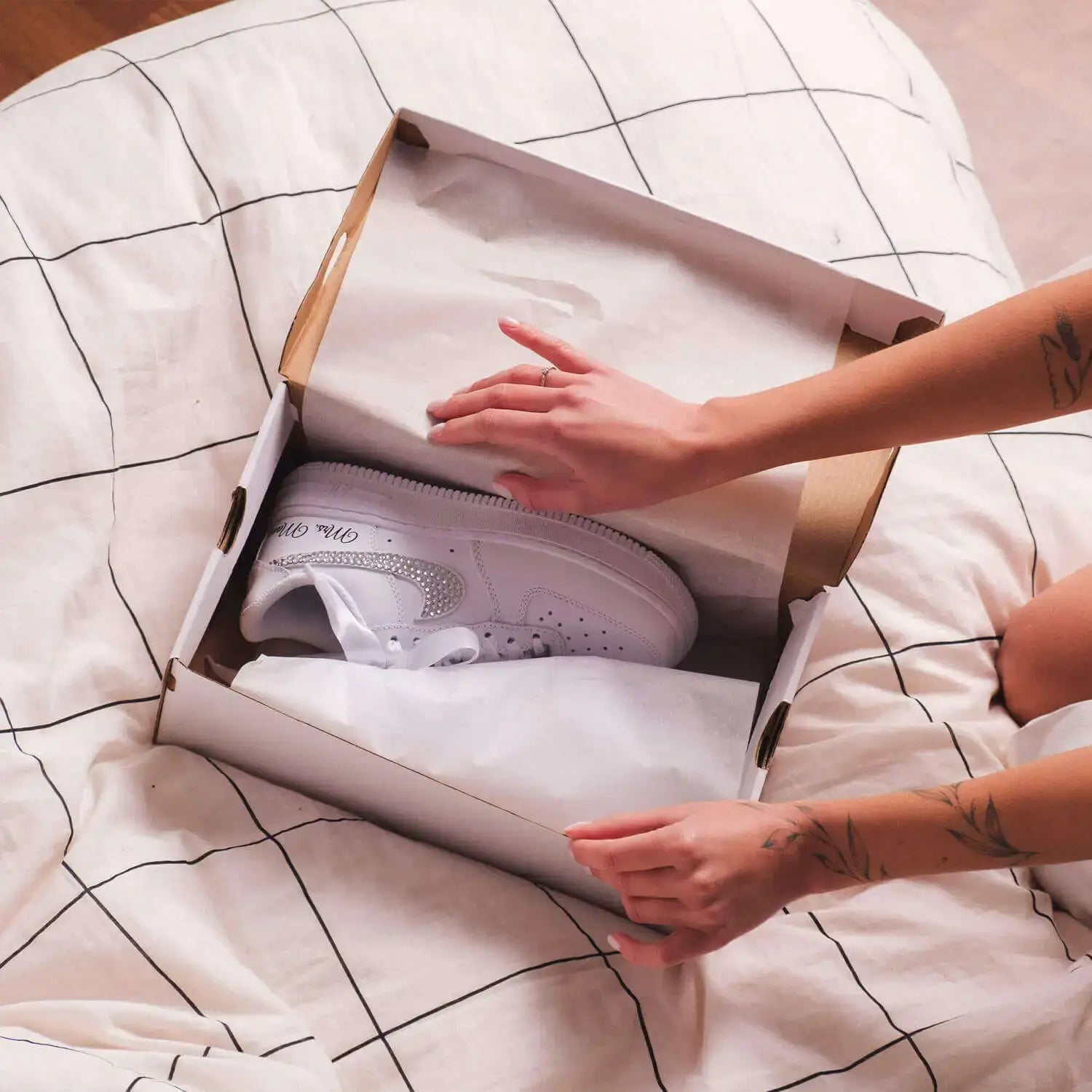 Person reaching into a box to grab a white sneaker on a bed with a grid pattern.