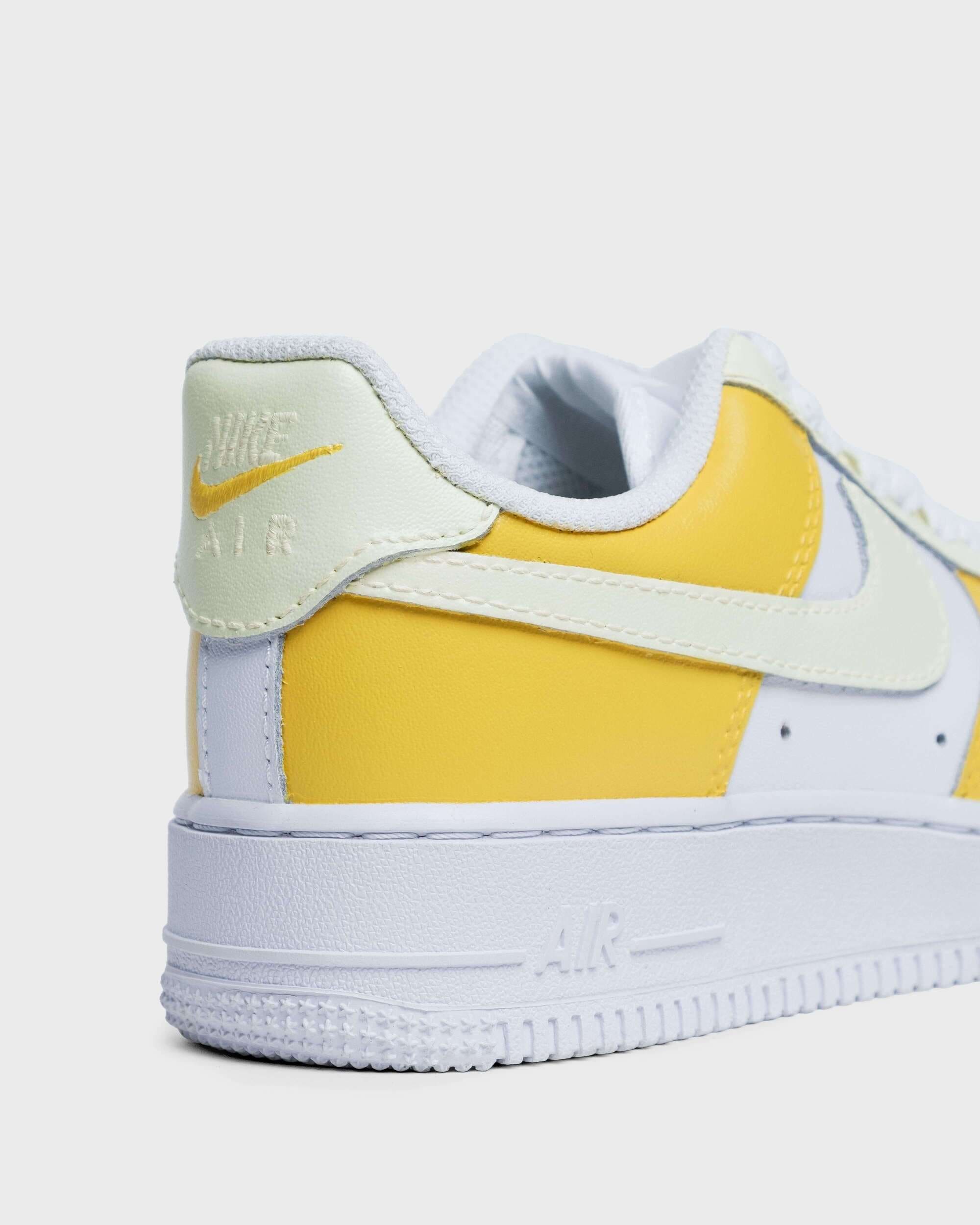 Close-up of sneaker: Close-up of a yellow and white custom Air Force 1 with a white Nike logo on a yellow background, showcasing the side of the shoe.