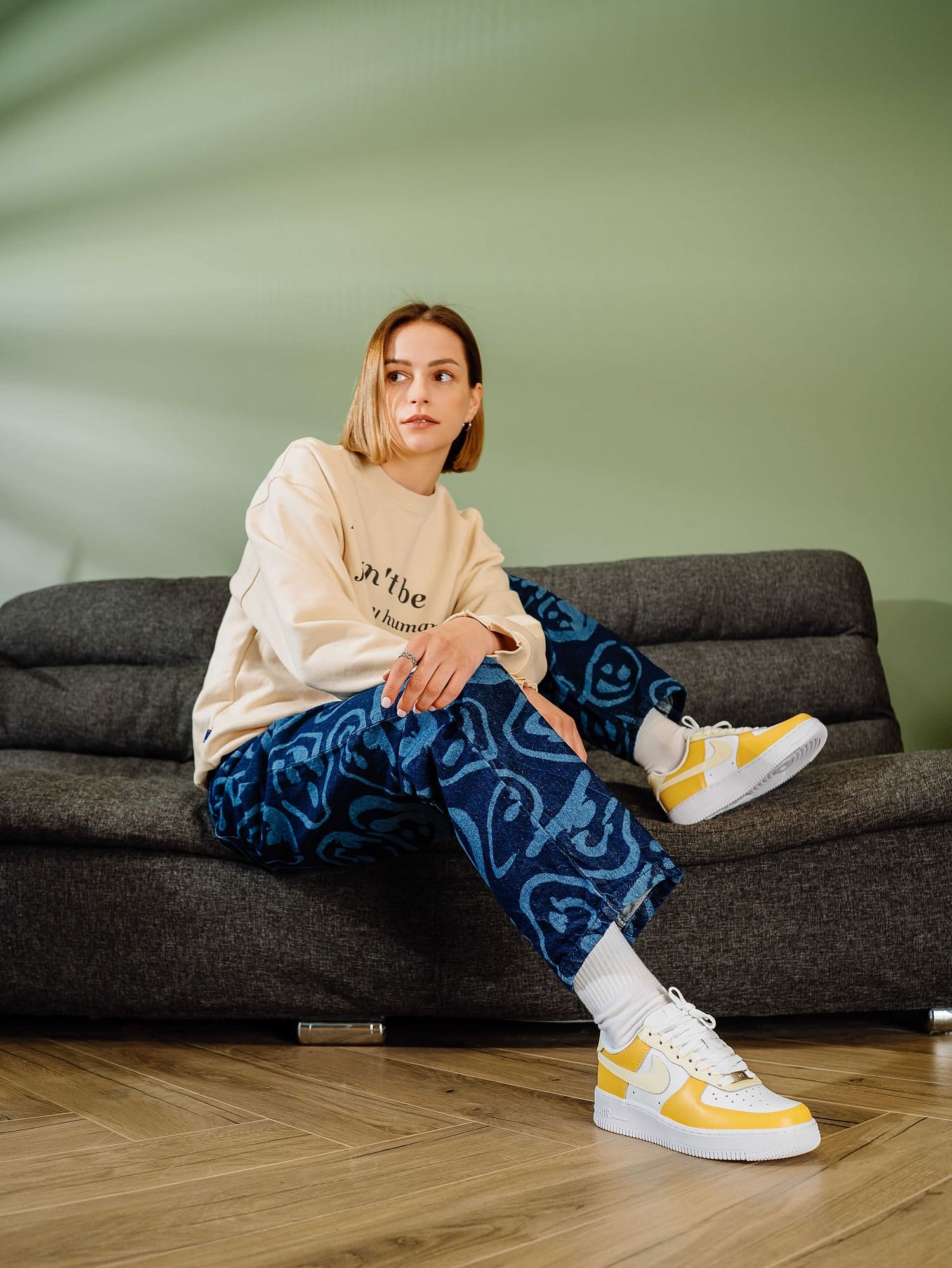 Model wearing sneakers while seated: Young woman sitting on a sofa wearing yellow and white custom Air Force 1 sneakers, showcasing the shoes styled with fashionable clothing.