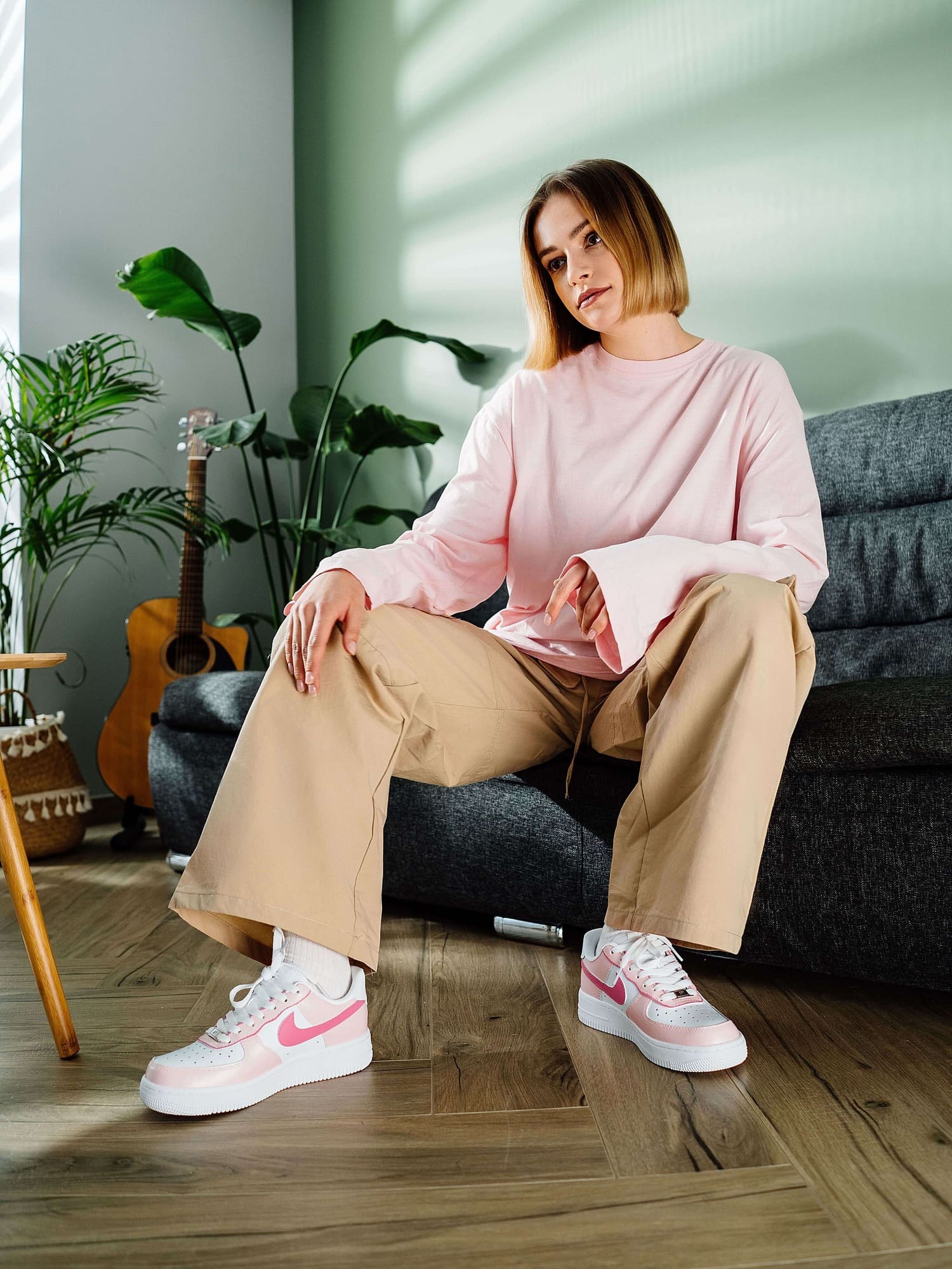 Model sitting on a couch wearing custom Air Force 1 sneakers with a light pink swoosh, paired with a casual beige and pink outfit, emphasizing a trendy, personalized look.