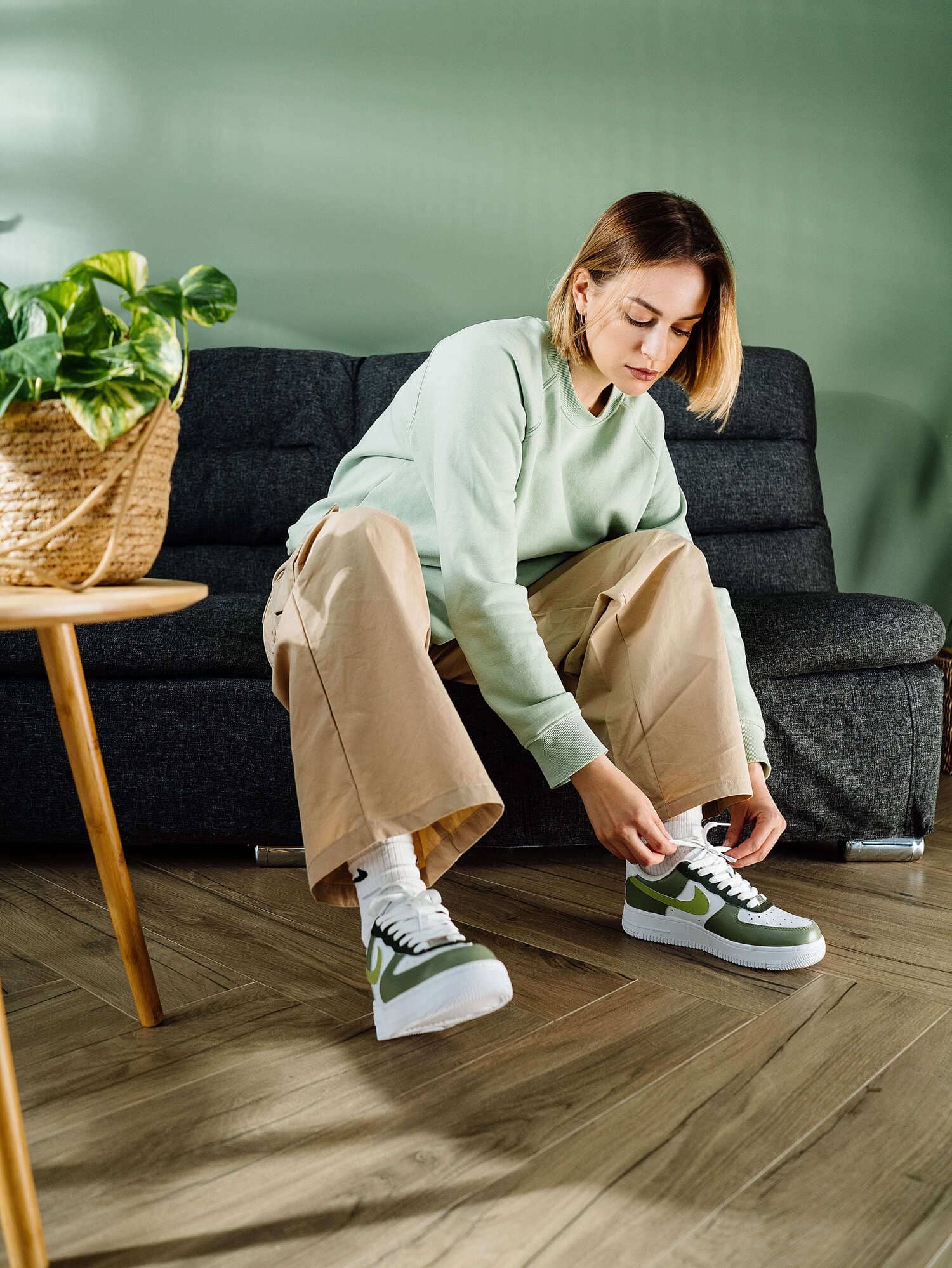 Person sitting on a sofa tying green Nike Air Force 1 sneakers with lime swoosh, wearing beige pants.