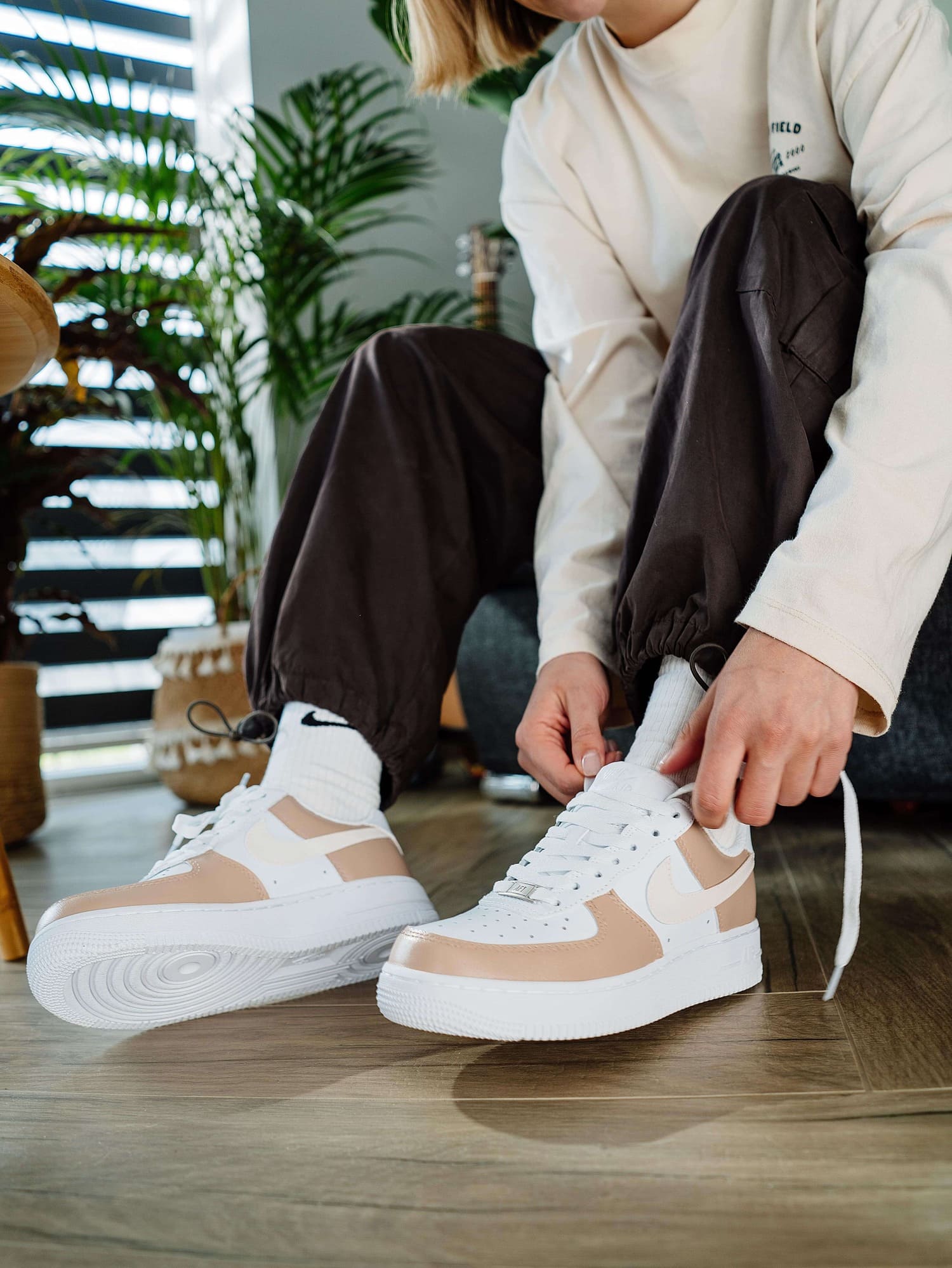 Person lacing up hand-painted custom Air Force 1 sneakers in cream tones, in a stylish indoor setting with plants.