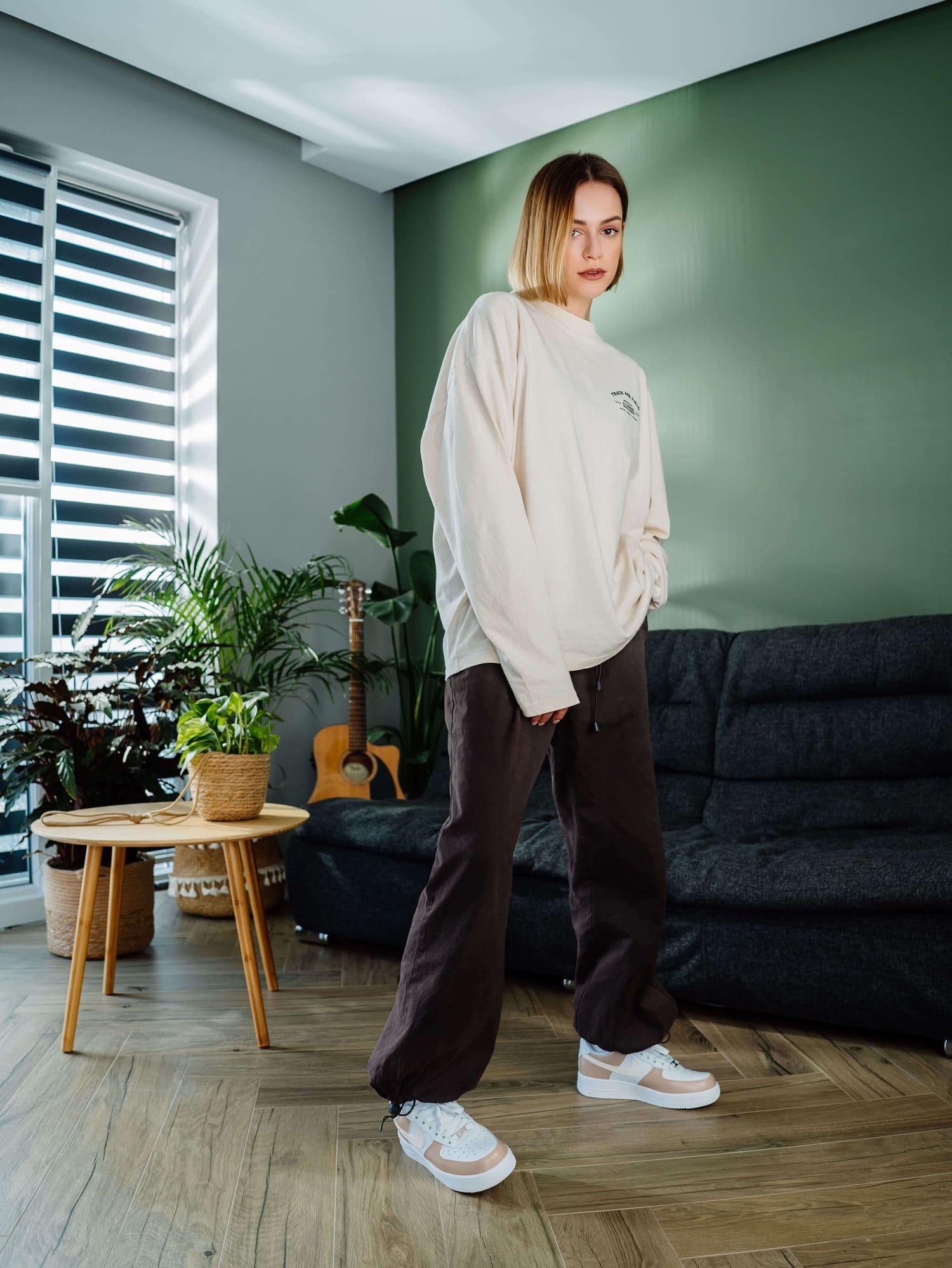 Model wearing hand-painted custom Air Force 1 sneakers in cream tones, standing in a modern living room with green accents