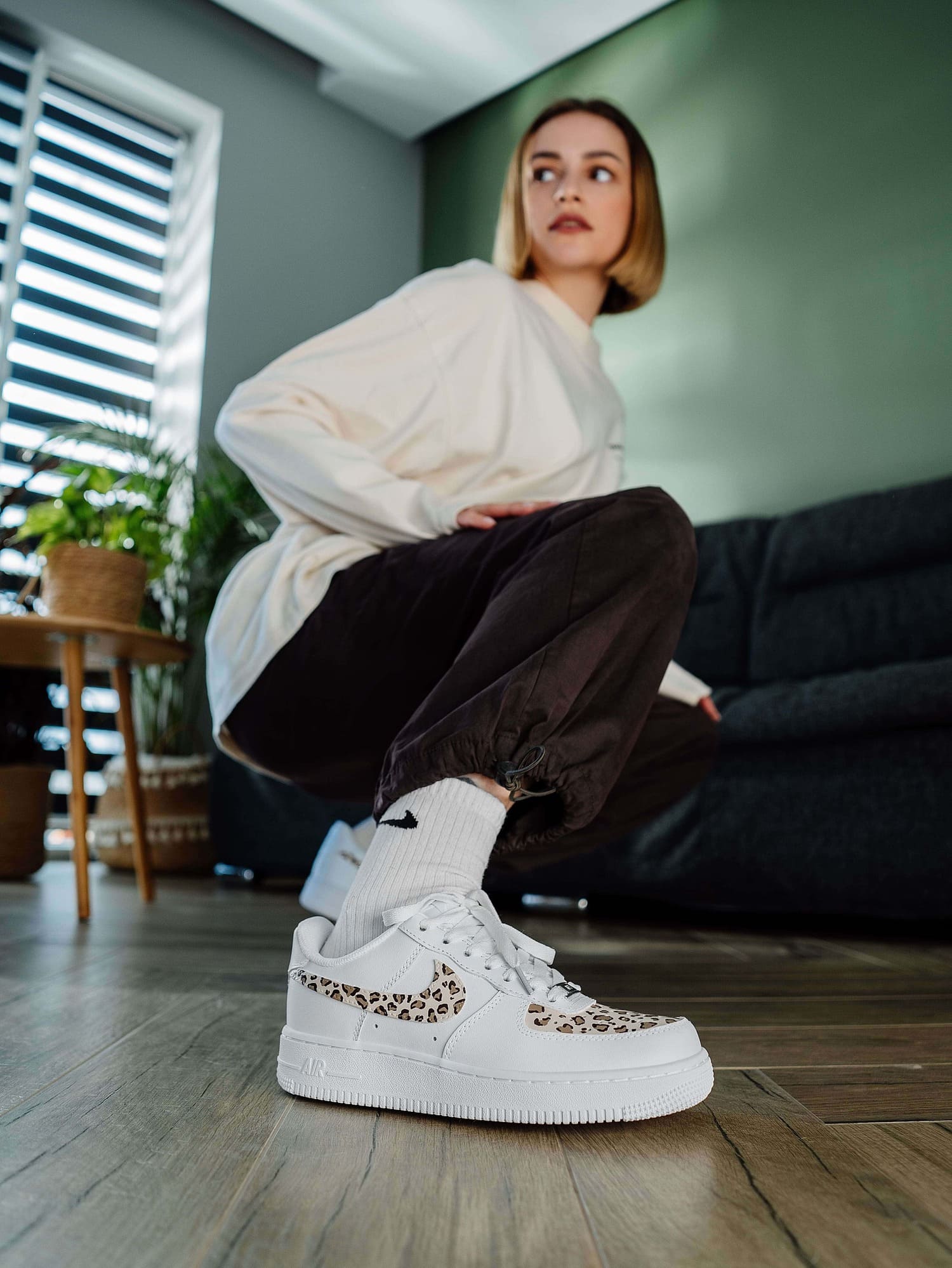 Model crouched down, showing custom Air Force 1 sneakers with leopard print on the swoosh and toe box, paired with relaxed-fit pants, emphasizing a stylish, personalized footwear choice.