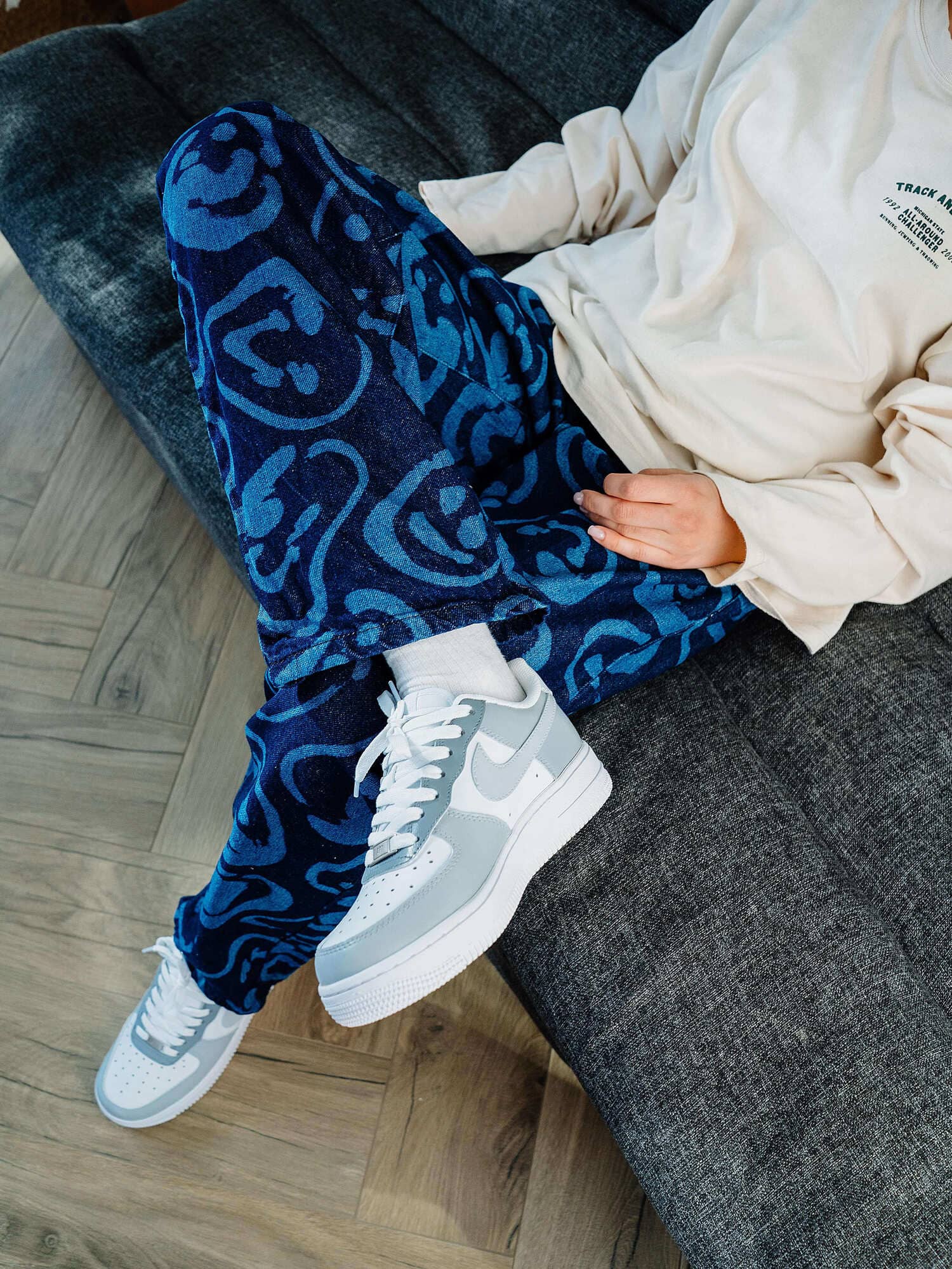 Model seated, wearing custom Air Force 1 sneakers with gray overlays, paired with blue patterned pants, highlighting a stylish, personalized footwear choice.