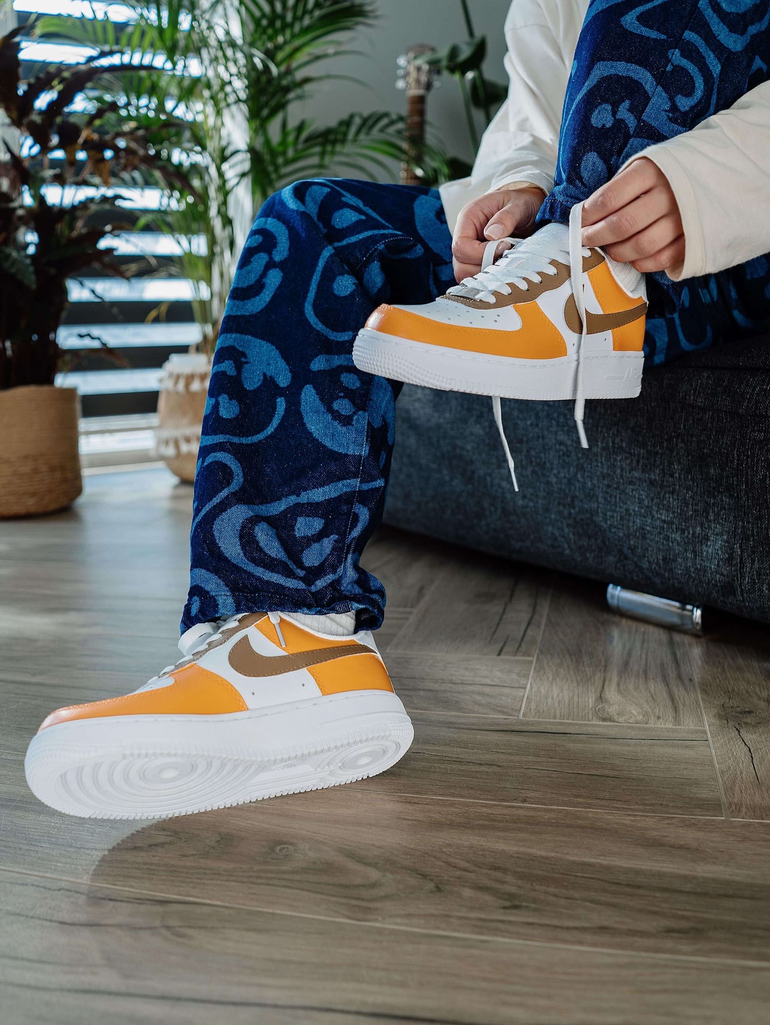 Model tying laces on custom orange and brown Air Force 1 sneakers, styled with patterned blue jeans, showcasing a vibrant, trendy look.