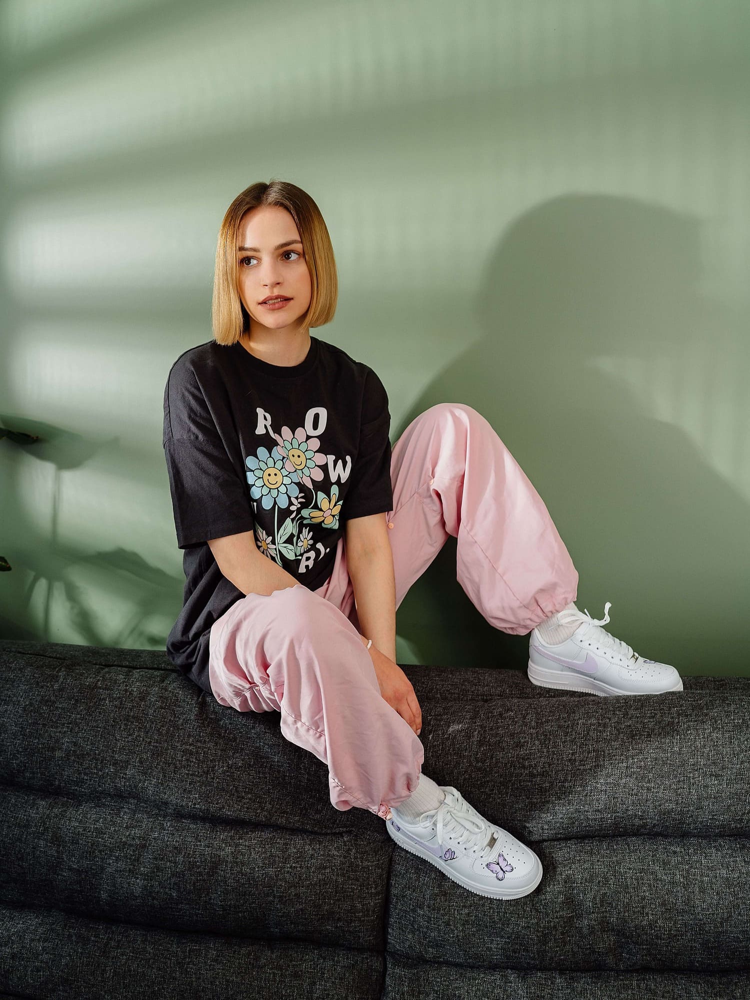 Model wearing hand-painted custom Air Force 1 sneakers with purple butterfly design, sitting on a modern couch in a stylish room. 