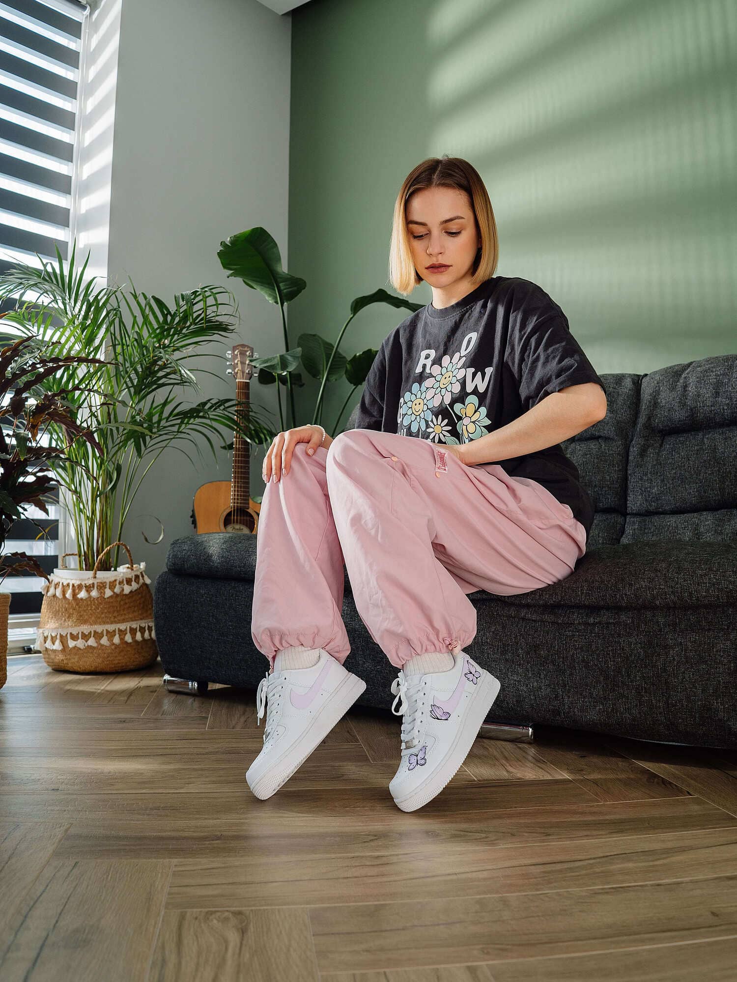 Model wearing custom Air Force 1 sneakers with purple butterfly design, sitting in a contemporary living room setting with plants