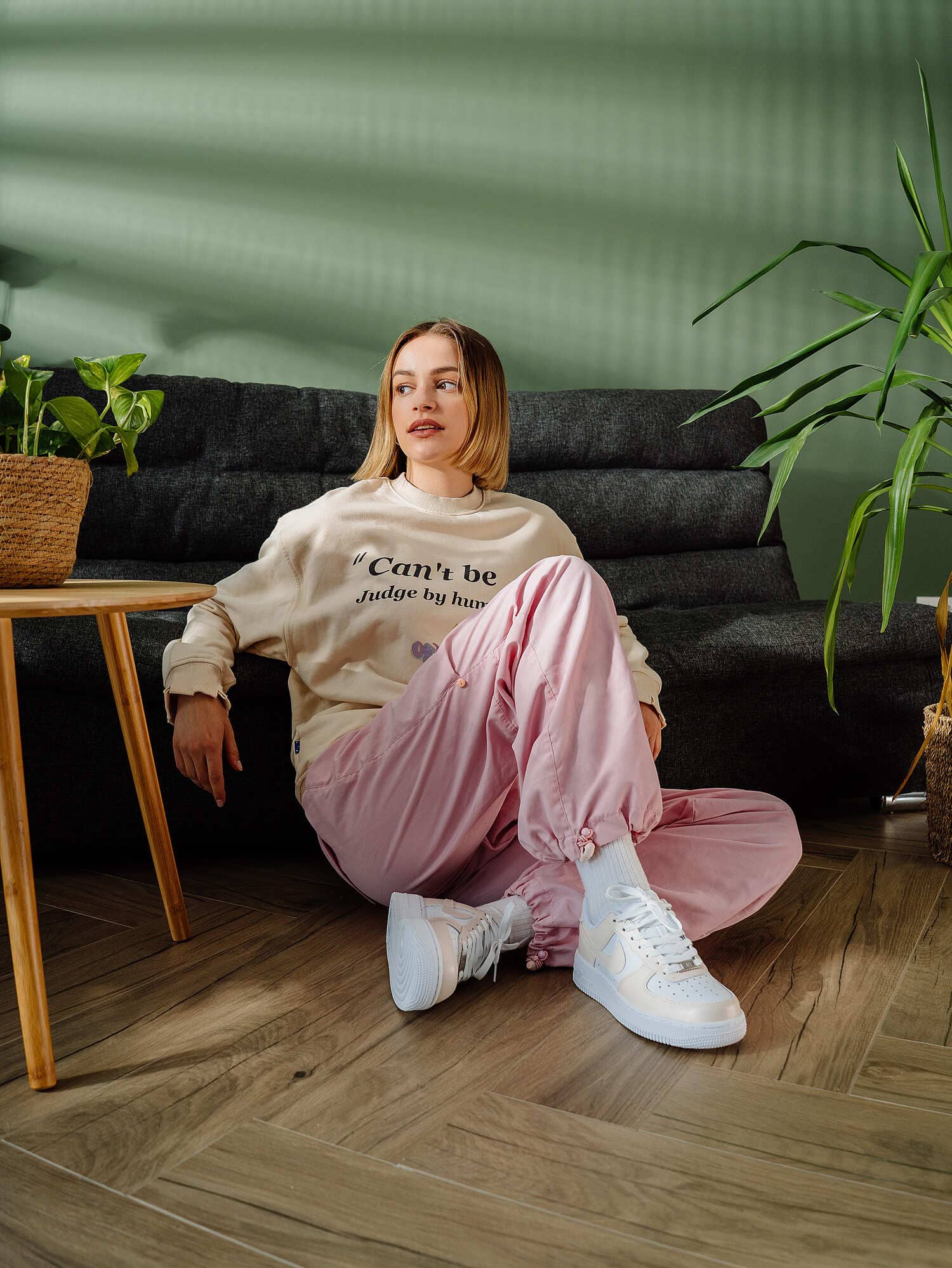 Model wearing custom Air Force 1 sneakers with a soft cream tone, sitting in a modern room with green accents.