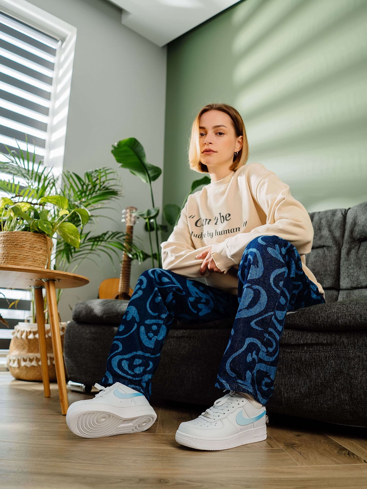 Model sitting on a couch wearing custom white Air Force 1 sneakers with light blue swoosh, paired with an oversized sweater and patterned jeans, creating a casual, stylish look.