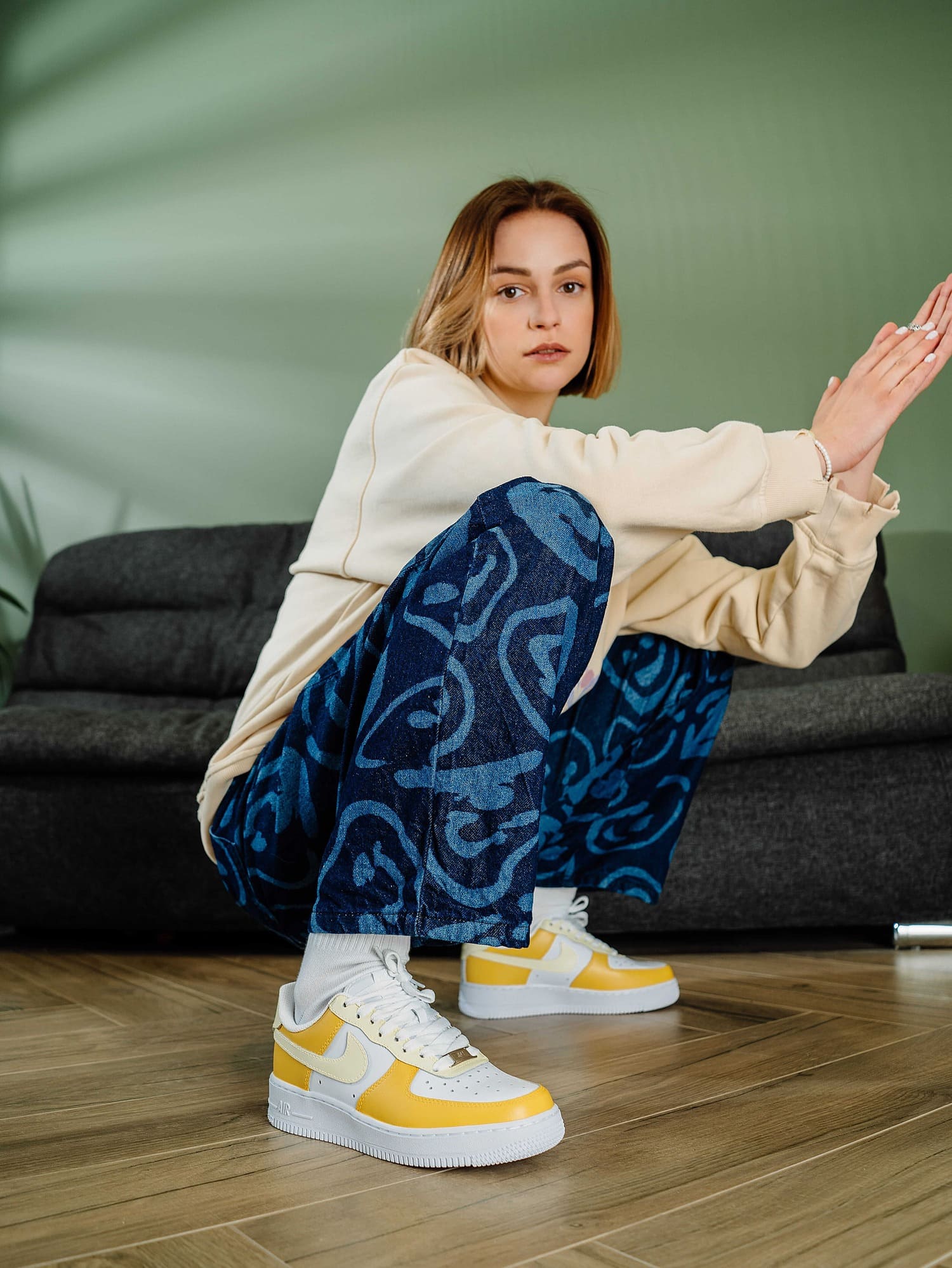 Model wearing sneakers in crouching pose: Young woman crouching on the floor in yellow and white custom Air Force 1 sneakers, displaying the shoes along with a trendy outfit.